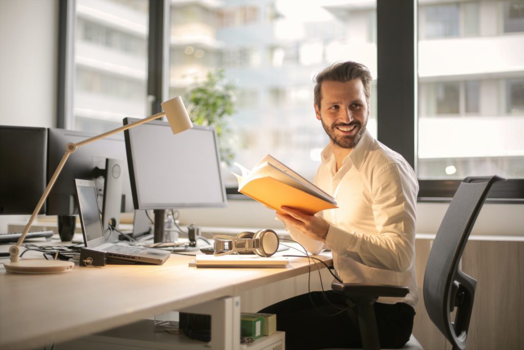 A man in a white shirt smiling while reading a book at a desk with multiple monitors and office equipment in a brightly lit room.