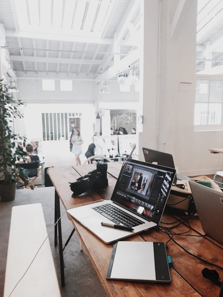 A laptop, tablet, and camera on a wooden table in a bright, spacious co-working space with people in the background.