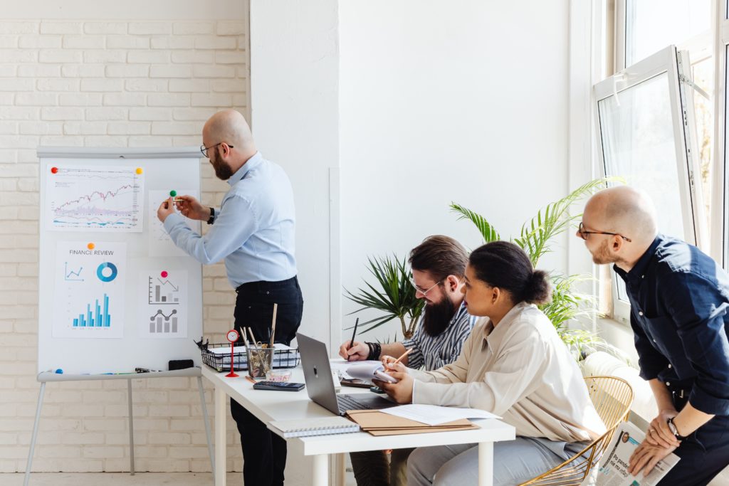 A business team discusses financial charts during a meeting as one man presents and three colleagues listen and take notes.