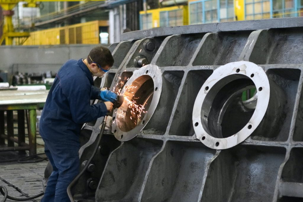 A worker in safety gear uses a welding tool on a large metal engine part in an industrial setting.