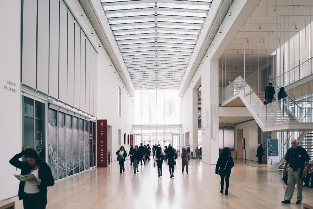 Interior of a modern art museum with visitors walking and reading brochures, featuring high ceilings and large windows.