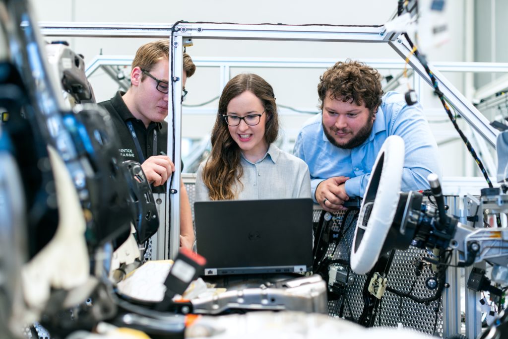Three engineers, one woman and two men, examine data on a laptop in a high-tech robotics lab.