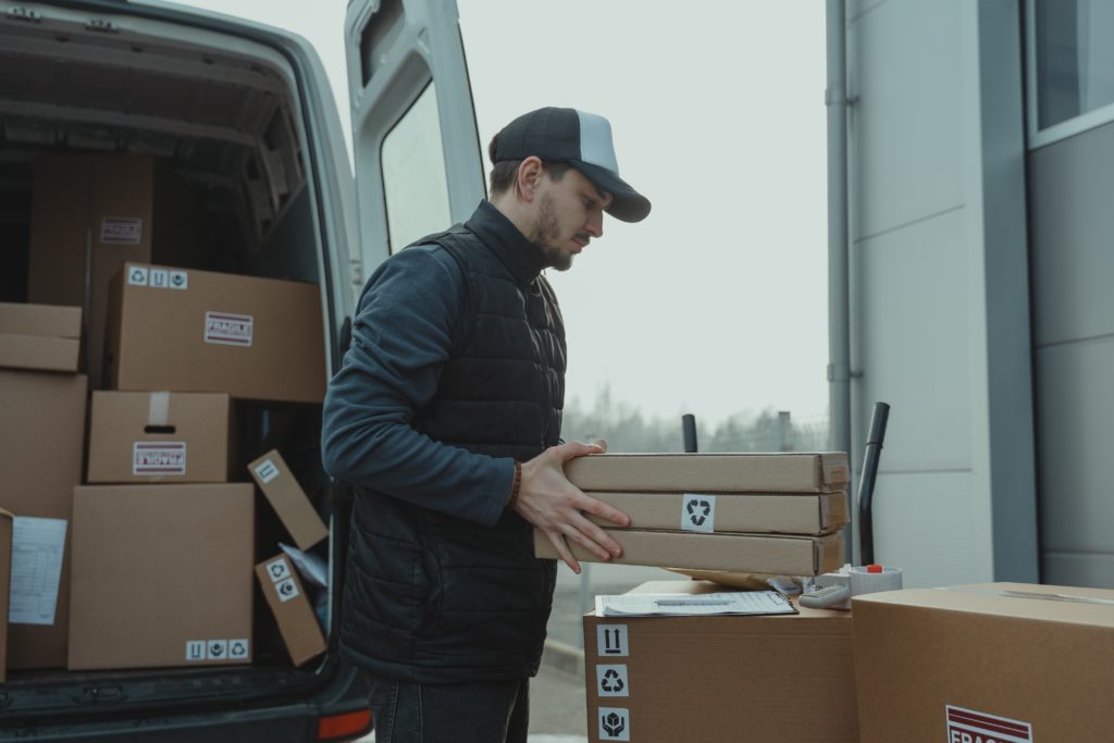 A delivery man in a cap and jacket unloads packages from a van outside a building.