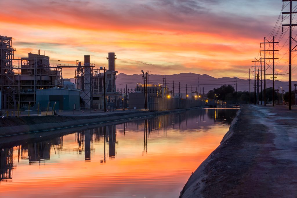 A vibrant sunset reflecting over a water canal beside an industrial facility with power lines and mountains in the background.