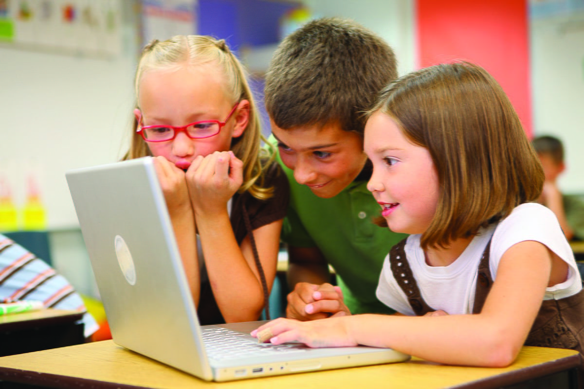 Three children, two girls and a boy, excitedly looking at a laptop screen in a classroom setting.