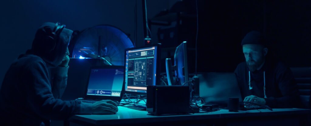Two people working on laptops in a dark room with blue lighting, focused on their screens with audio equipment visible.
