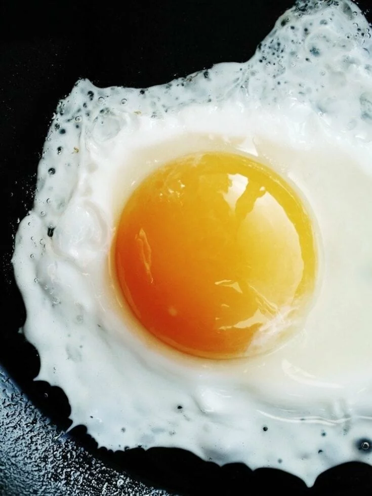A single sunny-side up egg frying in a black skillet, with the yolk intact and the whites bubbling around it.
