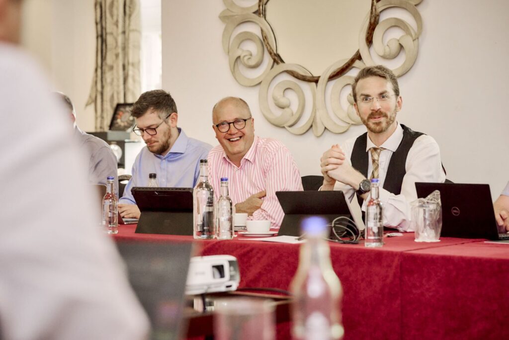Business professionals in a meeting room with laptops, focusing on a discussion; one man in a striped shirt is laughing.