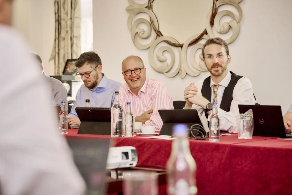 Business professionals in a meeting room with laptops, focusing on a discussion; one man in a striped shirt is laughing.