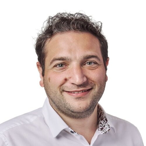 A portrait of a smiling man with curly hair and a light beard, wearing a white shirt with a floral collar.