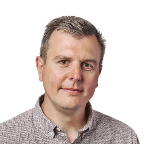 Portrait of a middle-aged man with short gray hair and fair skin, wearing a checkered shirt, looking at the camera with a subtle smile.