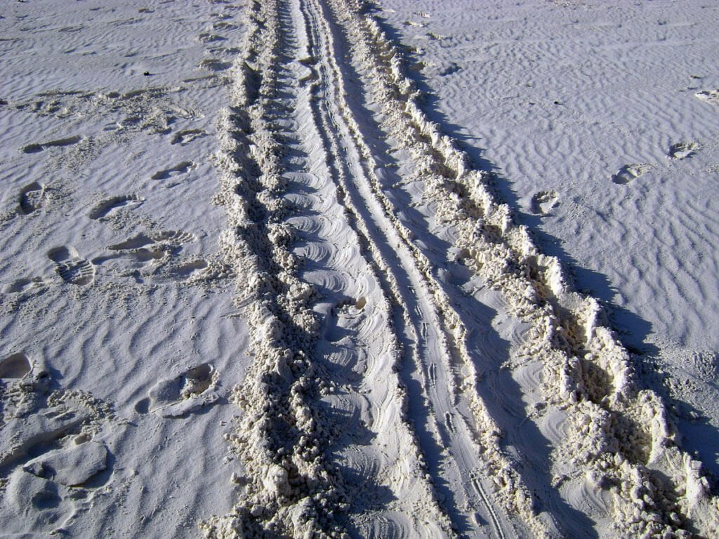 Tire tracks and footprints in the sand, showcasing a mix of deep vehicle ruts and scattered human steps under bright sunlight.
