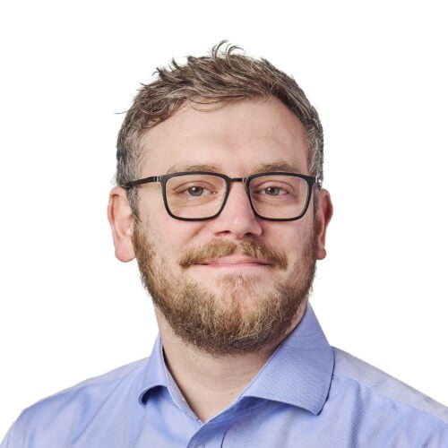 Portrait of a smiling man with a beard and glasses, wearing a blue shirt against a white background.