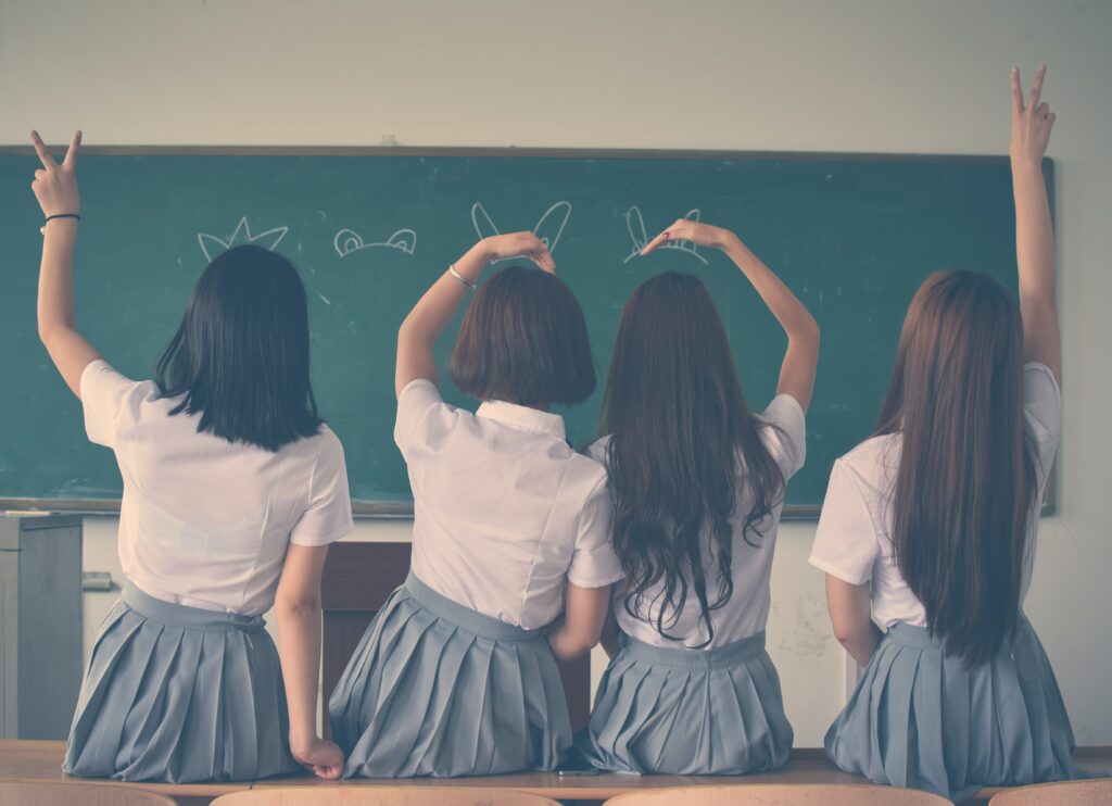 Four girls in school uniforms sit at a desk, drawing on a chalkboard in a classroom.