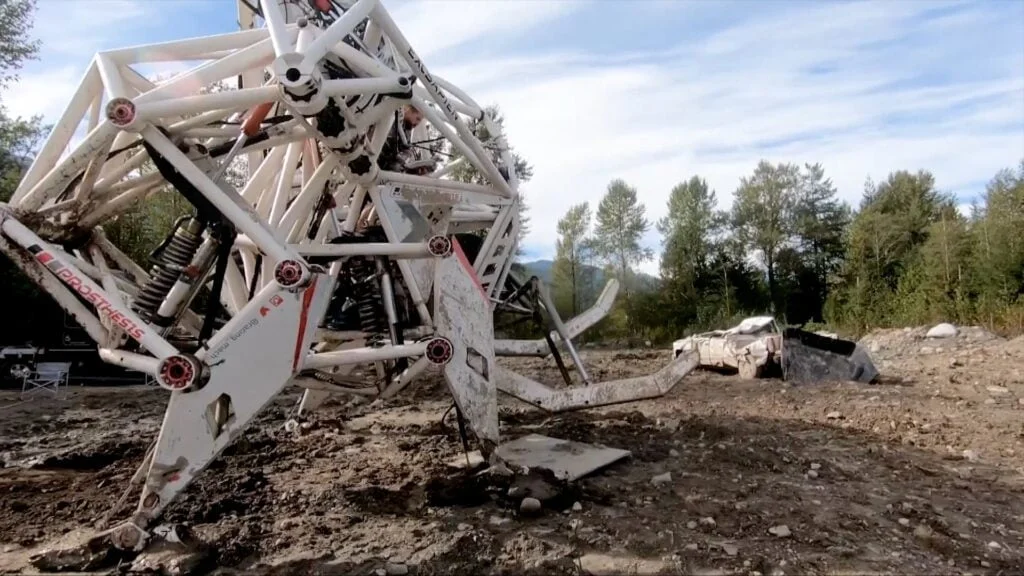 A large, multi-legged robotic structure stands on a barren terrain with trees and mountains in the background.