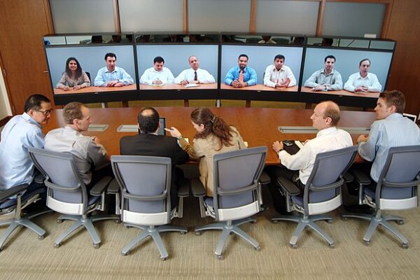 A group of professionals sit around a table in a conference room, engaging with multiple colleagues displayed on large video screens.