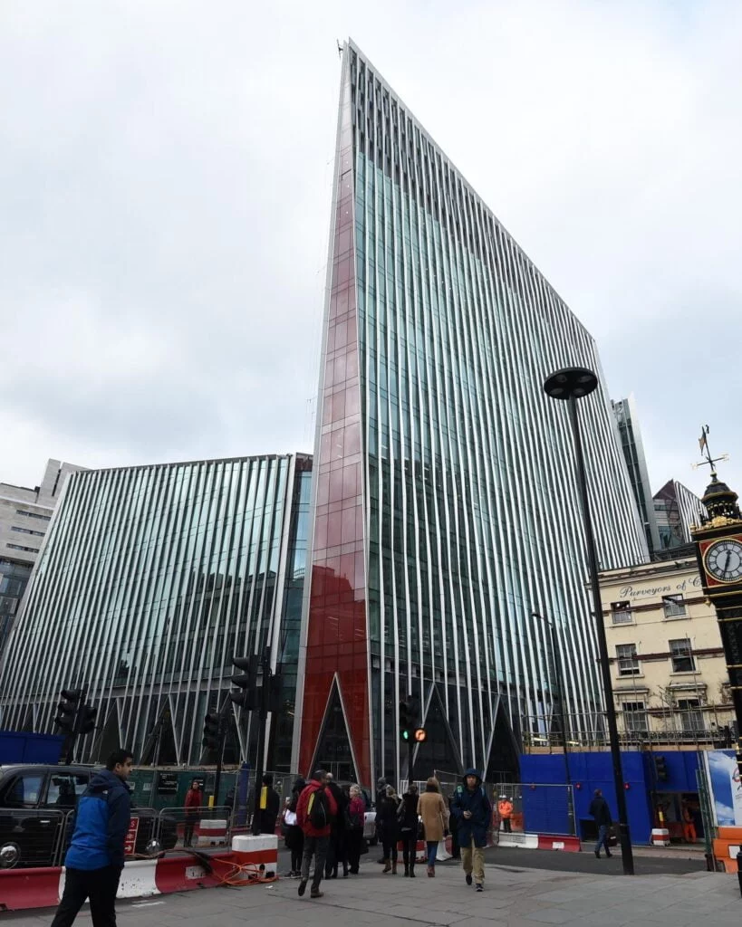 Modern glass building with a sharp, angular design, situated in an urban setting with pedestrians and street activity visible.