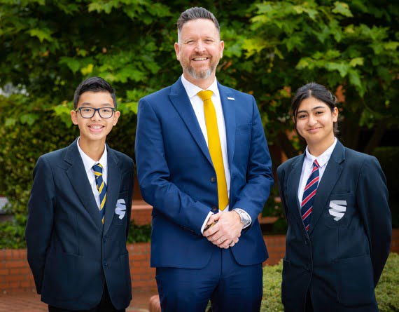 Two students and a teacher, all smiling and dressed in school uniforms, stand together outdoors discussing PRINCE2 project management.