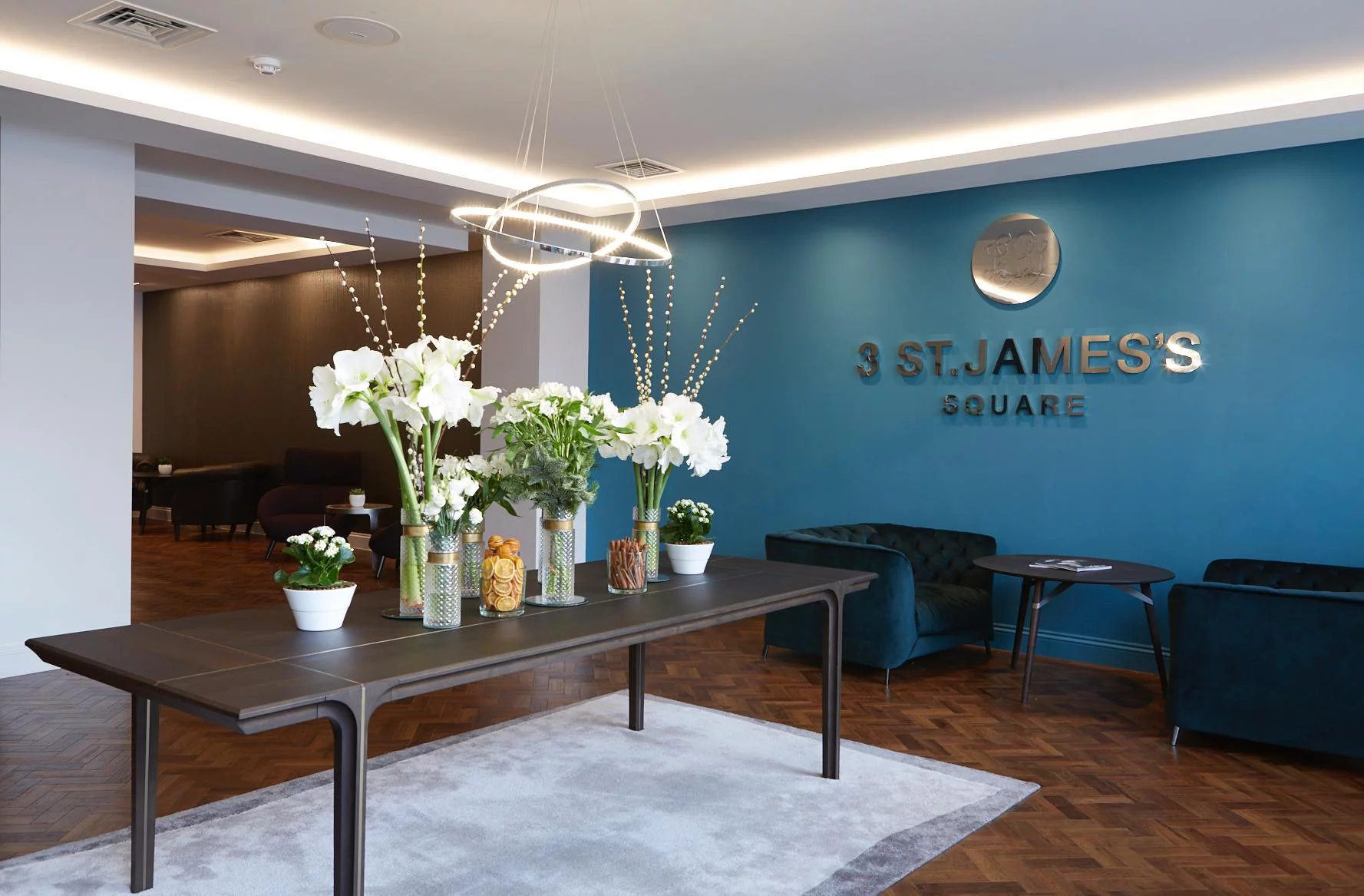 Modern office lobby with a teal accent wall featuring the text "3 st. james’s square", stylish furniture, floral arrangements, and contemporary lighting connected to the Internet of Things.