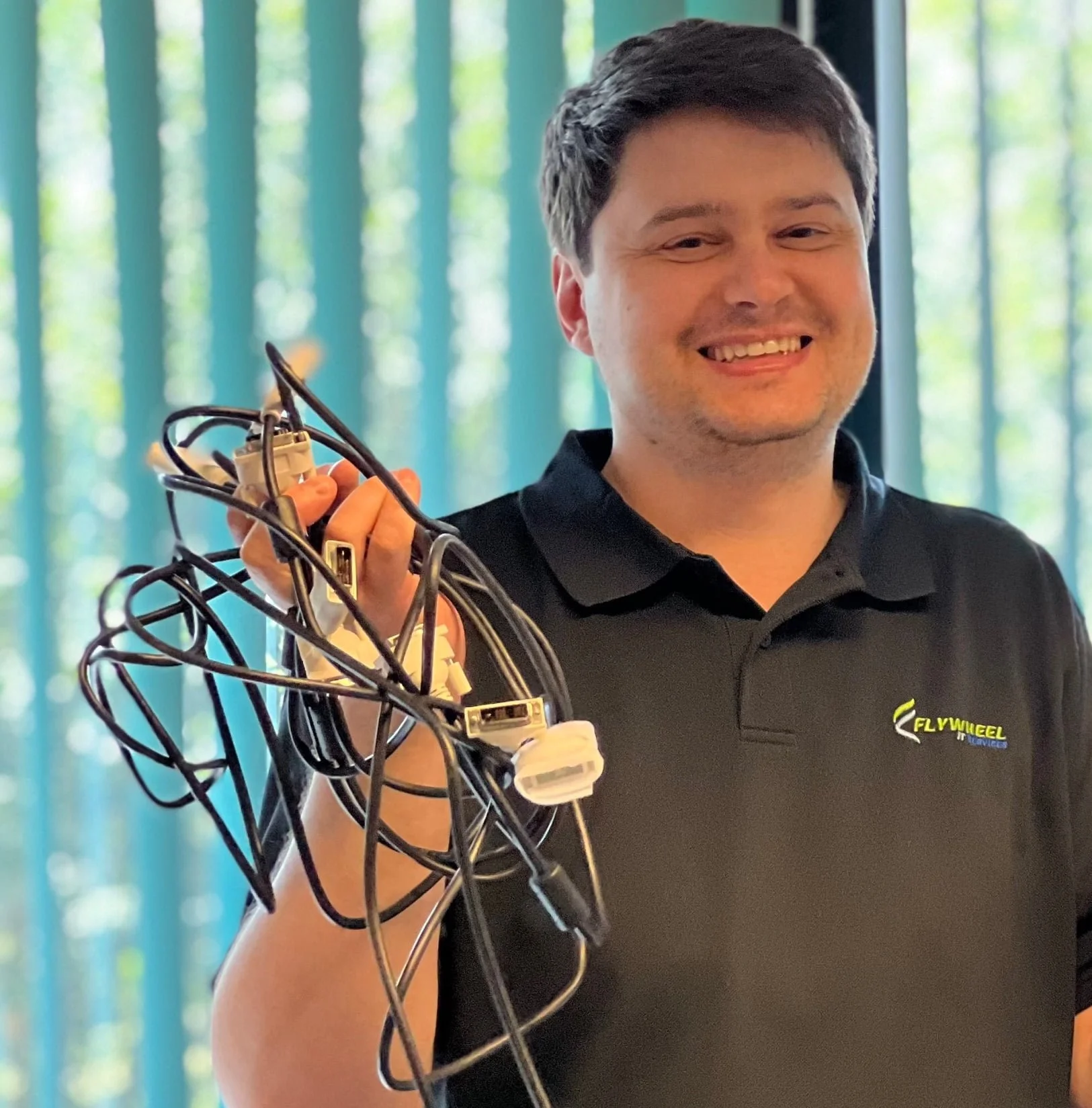 A man in a black polo shirt smiling and holding a tangled bunch of cables, providing IT services indoors.