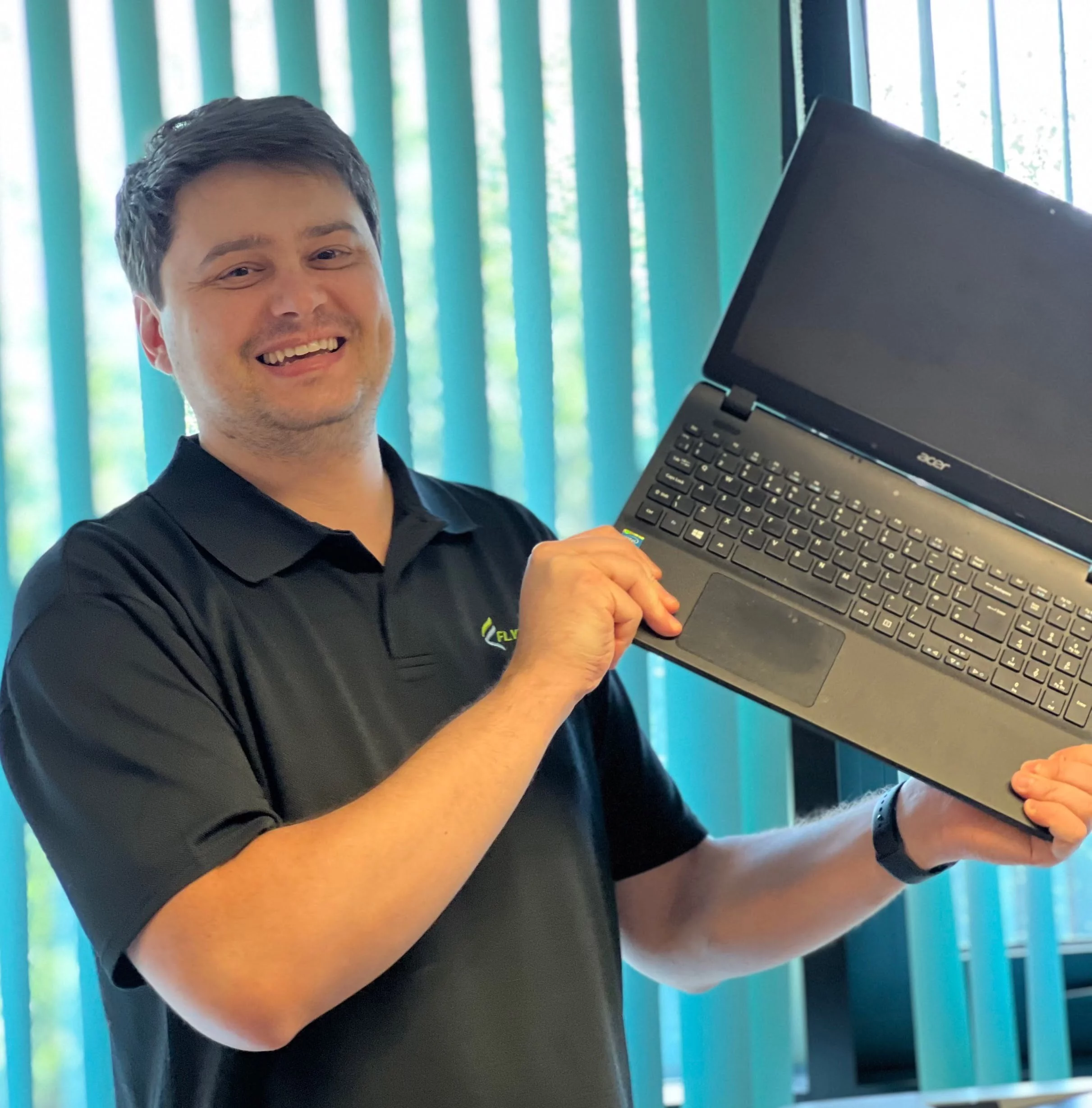 A smiling man in a black polo shirt holds an open laptop featuring ICT services for schools in an office, with vertical window blinds in the background.