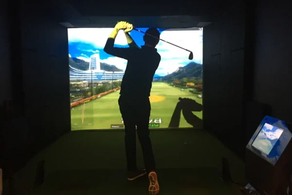 Man playing golf on an Internet of Things-enabled indoor simulator, standing on a green mat and swinging towards a large projected image of a golf course.