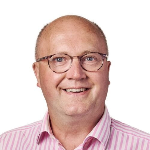 Meet the team: Portrait of a smiling bald man wearing glasses and a pink striped shirt against a white background.