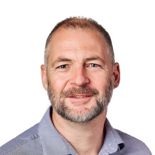 Meet the team: Middle-aged man with a beard, smiling at the camera, wearing a blue checked shirt against a white background.