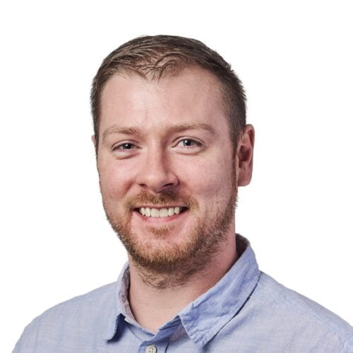 Meet the team: Portrait of a smiling man with light brown hair and a beard, wearing a blue button-up shirt against a white background.