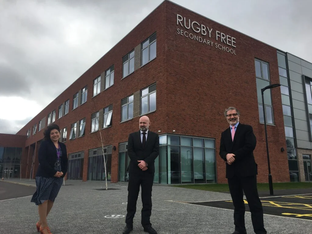 Three individuals standing in front of the rugby free secondary school complex, a modern brick building, under a cloudy sky.