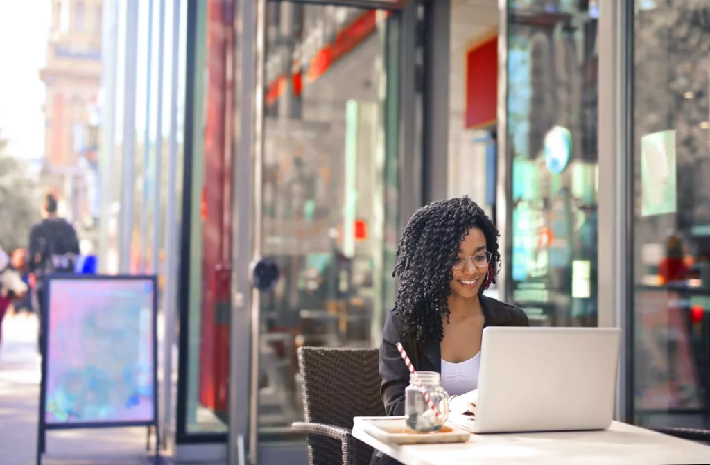 A young woman with curly hair smiles while using a laptop to access IT services at an outdoor café table on a sunny city street.