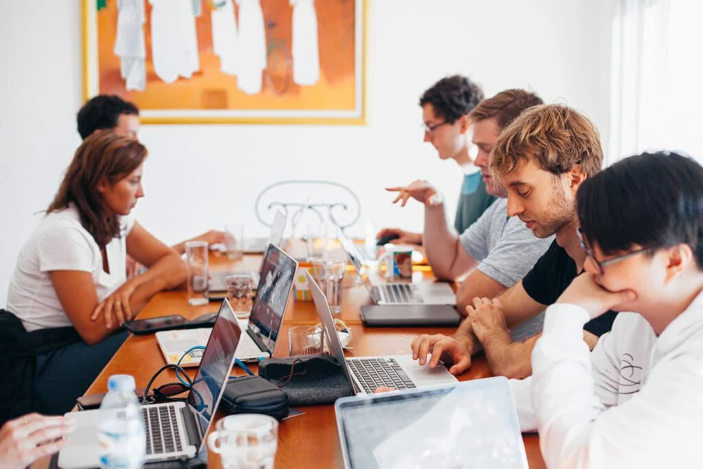 A group of six IT services professionals, engaged in a meeting around a table filled with laptops, in a brightly lit office space.