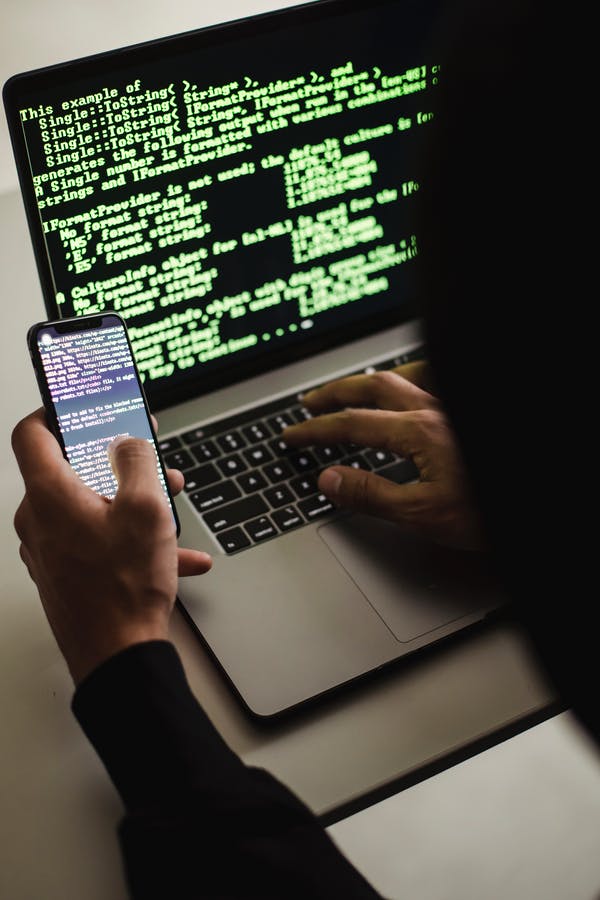 A person coding on a laptop while also holding and looking at a smartphone, both screens displaying lines of code for their Cyber Essentials Certification project.