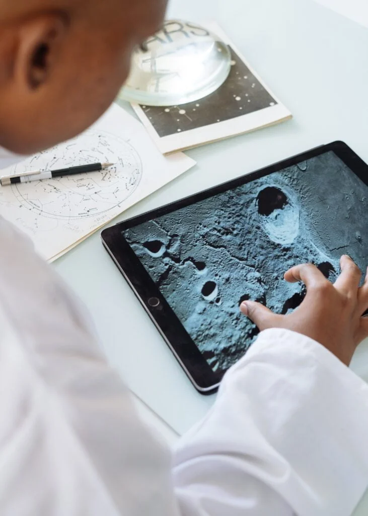 Scientist examining a high-resolution image of the moon's surface on a tablet, with notes and IT services documentation on the desk.
