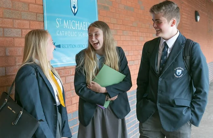 Three students in uniforms laughing together outside st michael's catholic school, near the reception office sign.