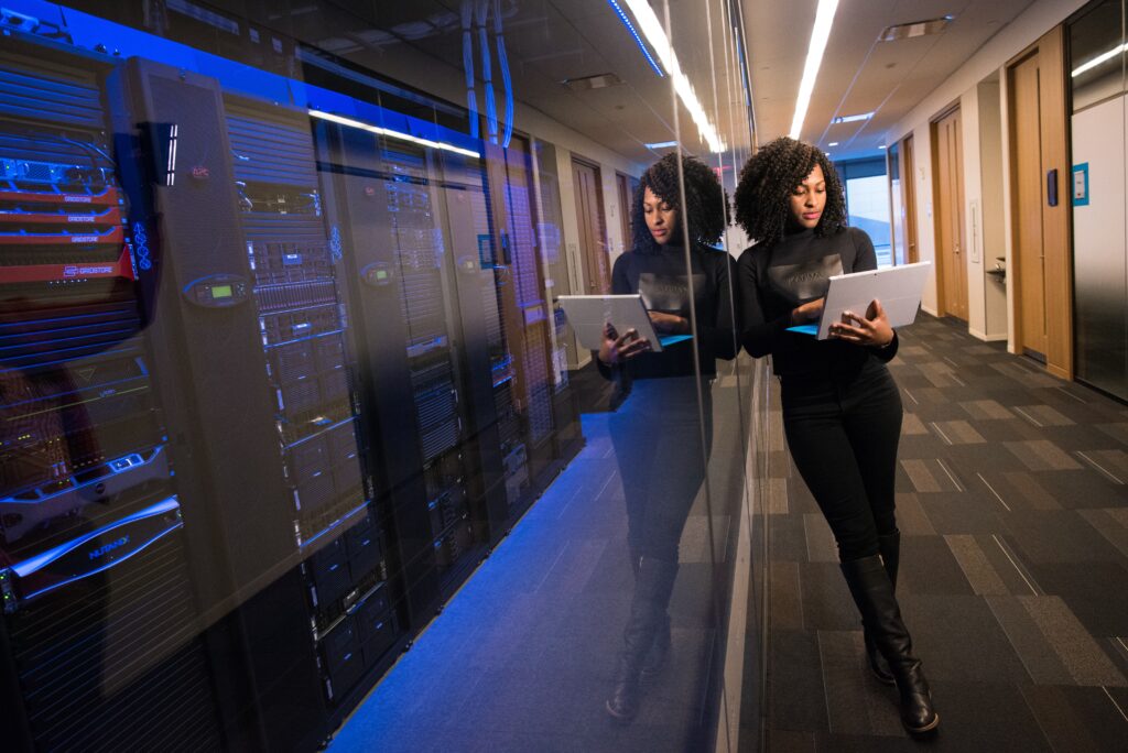 Two women with tablets discuss outsourced IT support in a server room with illuminated racks of data equipment.