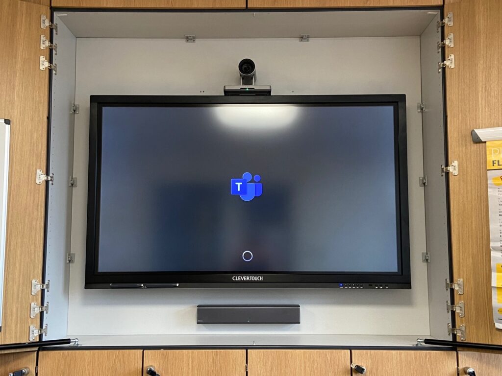 A wall-mounted clevertouch screen, part of the audio visual systems, displaying a blue and white logo inside a gray cabinet with open doors.