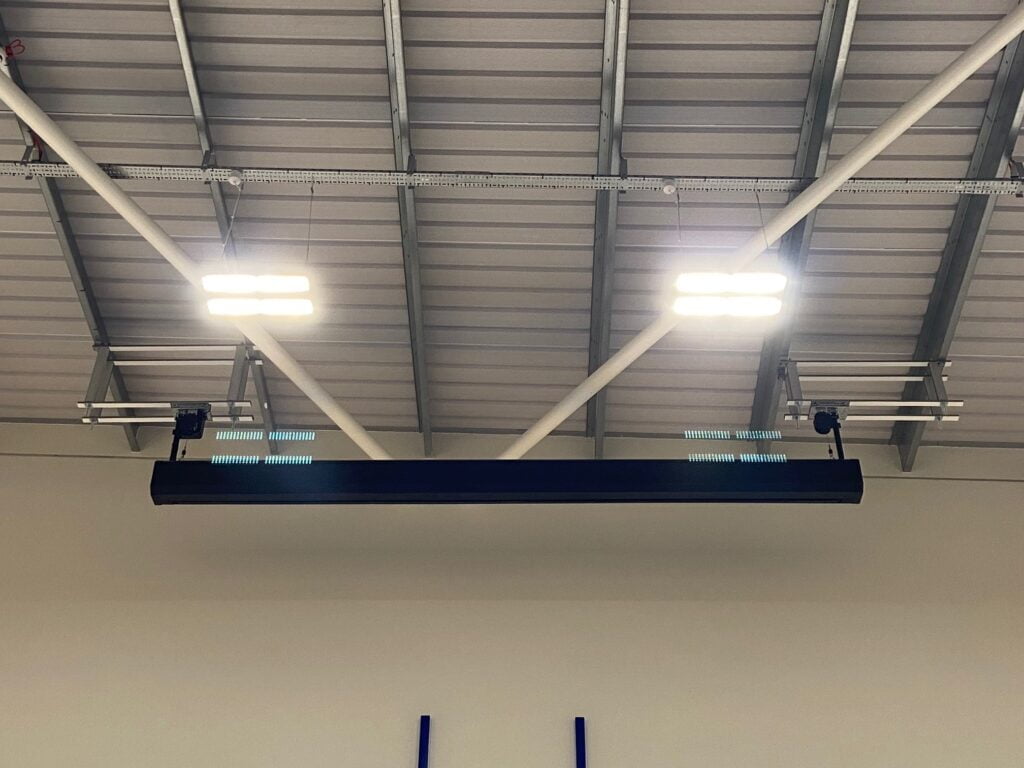 Ceiling-mounted industrial light fixtures in a warehouse, with visible steel beams and a video wall.