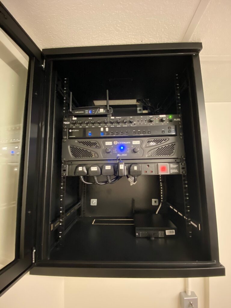 A wall-mounted server rack containing various networking equipment with visible cables and LED indicators near a video wall in a dimly lit room.