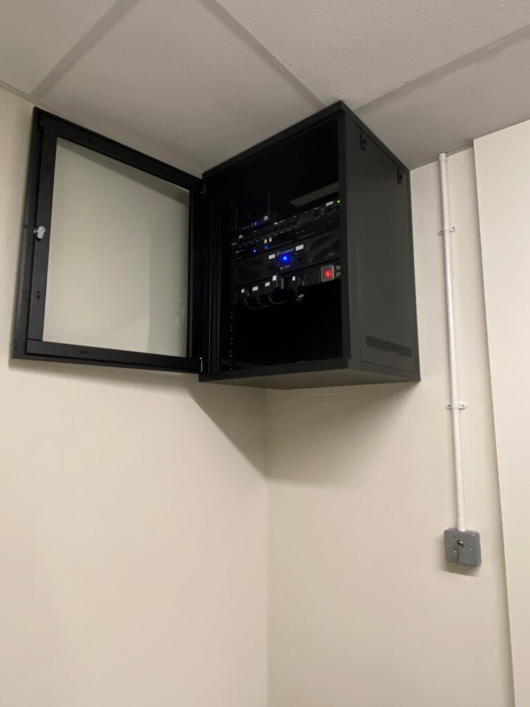 Open black cabinet on a wall containing network equipment with visible cables and LED indicators, next to a white conduit pipe and a video wall.