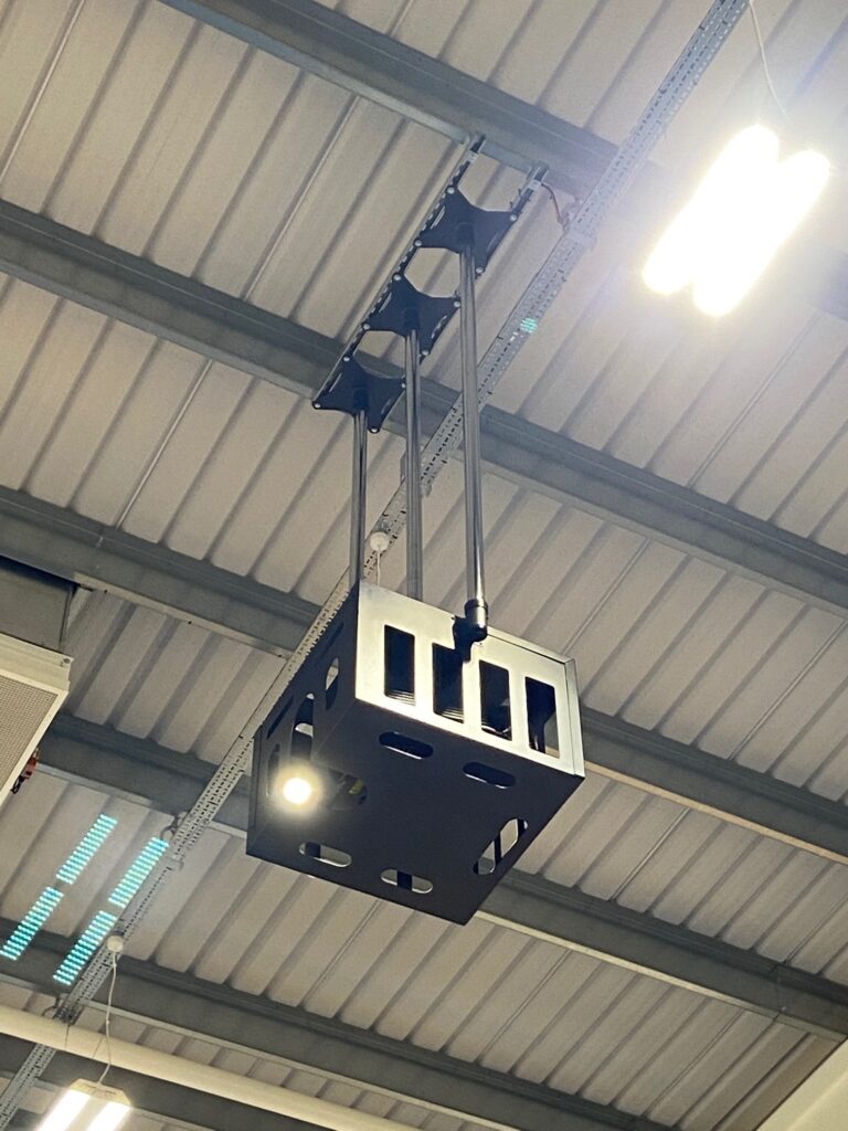 Industrial black crane hoist lifting a metal box in a warehouse, showing metal beams and video wall structure.
