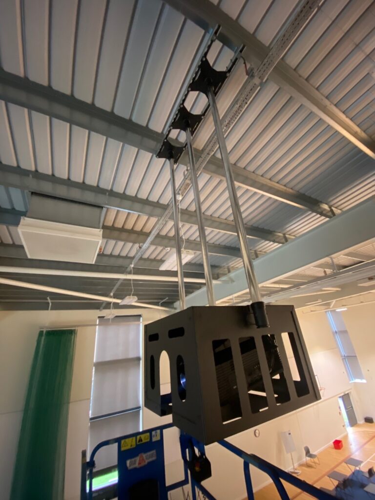 Ceiling-mounted video wall projector in a room with exposed metal beams and translucent window blinds.