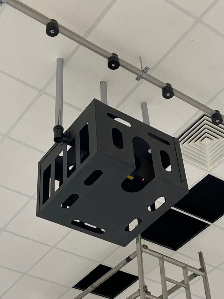 A black cubic structure with holes, possibly an audio visual systems projector mount, hanging from a white ceiling with visible ductwork and scaffold.
