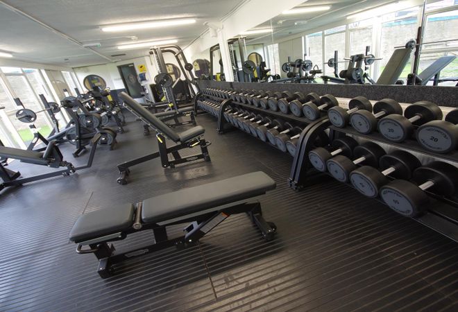 Interior of a leisure centre showing a row of dumbbells, weight benches, and various exercise machines, set against large windows.