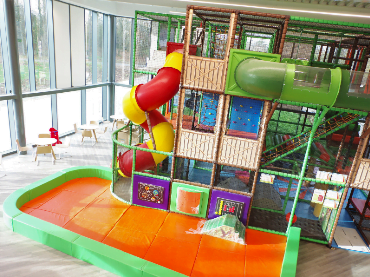 Indoor leisure centre with a colorful climbing structure and slide, surrounded by padded flooring and large windows.