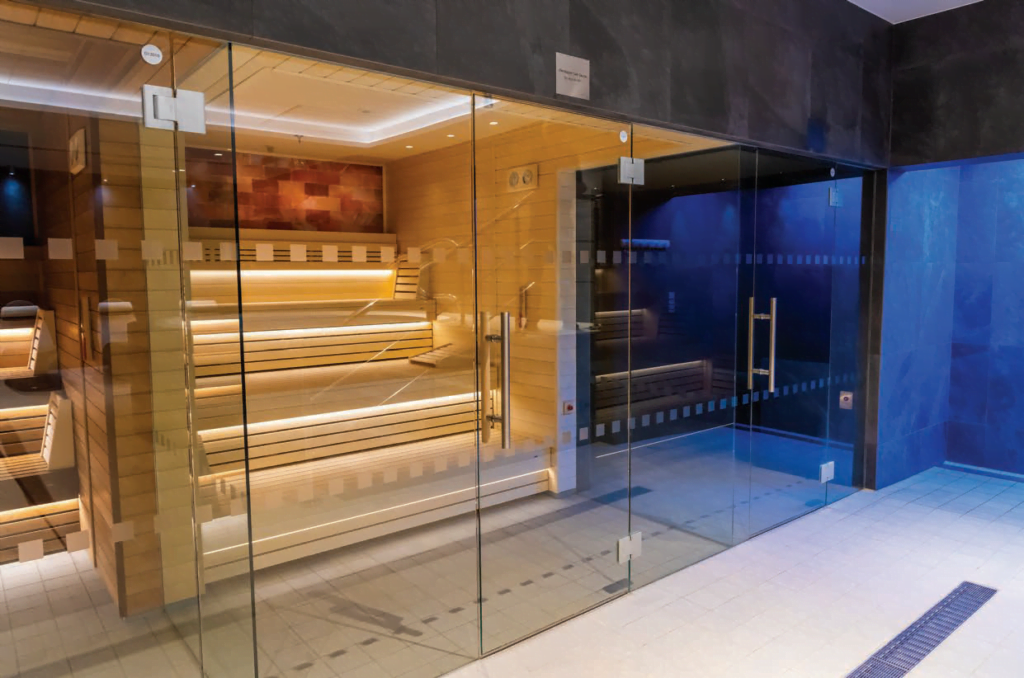 A modern sauna room in a leisure centre, featuring wooden benches and a glass door, with blue tile accents and ambient lighting.