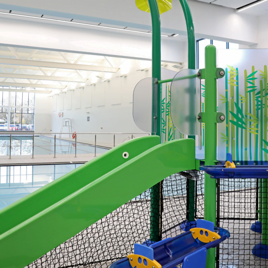 Indoor leisure centre equipment with a green slide and climbing structure next to a brightly lit swimming pool.
