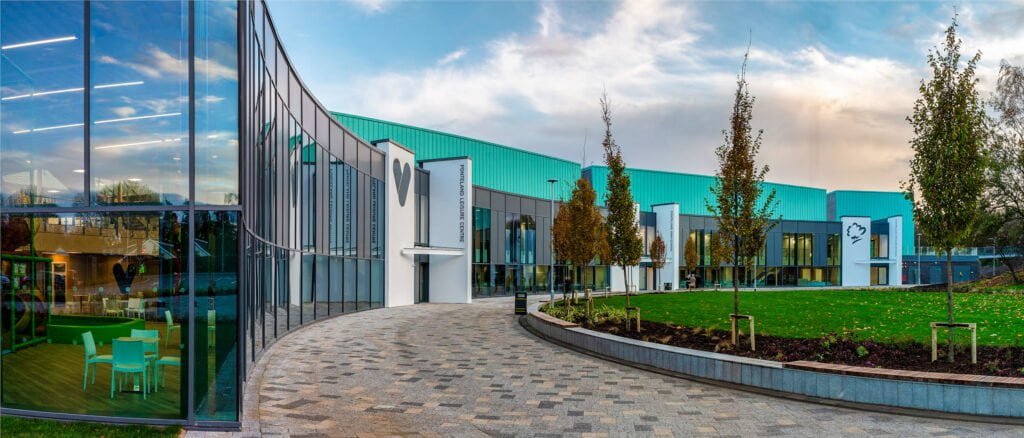 Panoramic view of a modern leisure centre with reflective glass windows and a curved pathway leading to its entrance, surrounded by green landscaping.