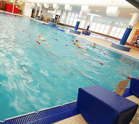 Indoor swimming pool at a leisure centre with swimmers in action, separated by lane ropes, viewed from a poolside angle with blue mats on the deck.