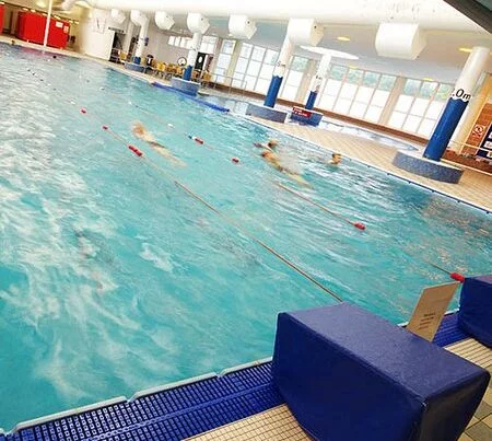 Indoor swimming pool at a leisure centre with swimmers in action, separated by lane ropes, viewed from a poolside angle with blue mats on the deck.